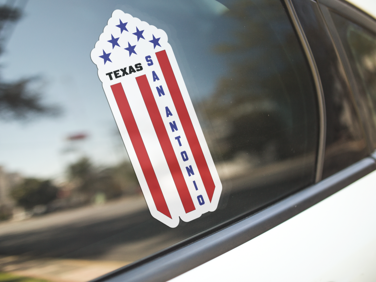 A die-cut vinyl sticker of the Texas San Antonio flag design, featuring red and white vertical stripes, blue stars, and the text 'SAN ANTONIO' in blue, shown applied to a car window.