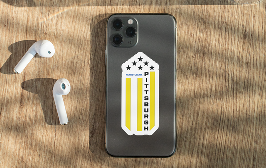 A dark gray smartphone case with a Pittsburgh flag design is shown on a wooden table next to white wireless earbuds. The case features a white shield with yellow stripes and the words 'Pittsburgh' and 'Pennsylvania'.