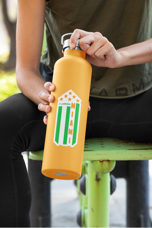 A woman sitting on an outdoor fitness bench holds a yellow water bottle with a green and white striped shield logo. She is wearing an olive green tank top and black leggings in a sunny park setting.